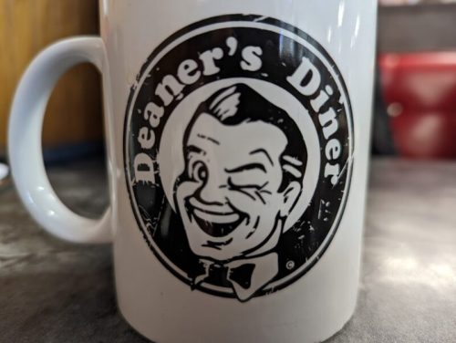 A close-up of a white coffee mug featuring a vintage logo of Deaner's Diner with a smiling, winking character.