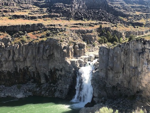 A waterfall cascades over rocky cliffs into a green pool, surrounded by rugged terrain and sparse vegetation.