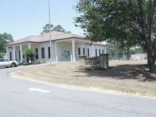 A city hall building with a sign, surrounded by trees and a parking area, on a sunny day.