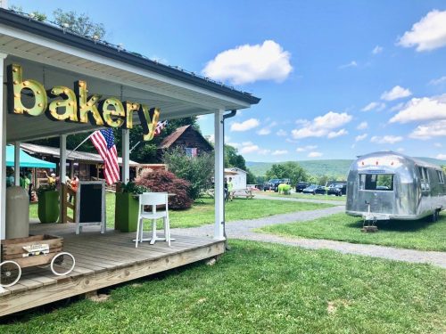 A charming bakery with a sign, surrounded by green grass and a vintage Airstream trailer under a blue sky.