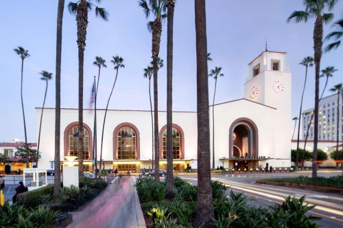 Historic train station with palm trees, clock tower, and evening lights, surrounded by lush greenery.