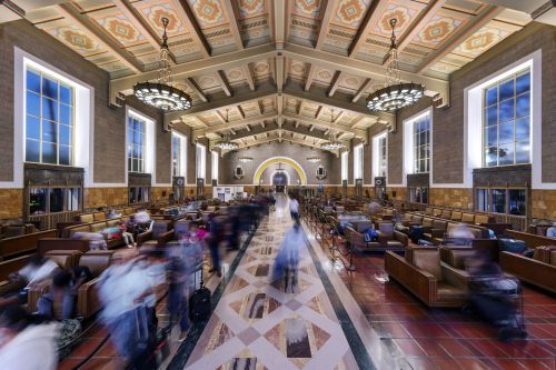 A bustling train station interior with high ceilings, large windows, and people moving through the space.