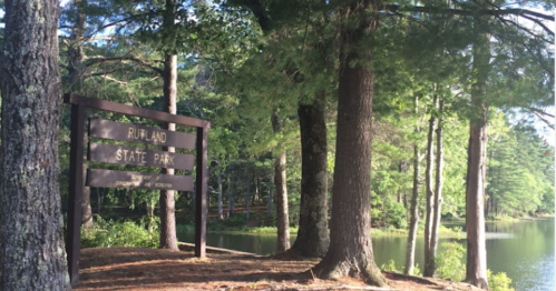 Sign for Rutland State Park beside a calm lake, surrounded by tall trees and greenery.