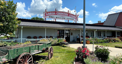 Amish Country Store with a welcome center sign, surrounded by flowers and a wooden wagon in front under a blue sky.