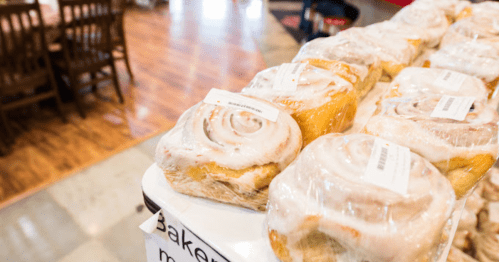 A display of cinnamon rolls wrapped in plastic, with a cozy bakery setting in the background.