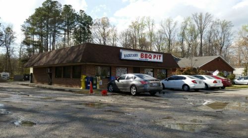 A brick building with a sign reading "Backyard BBQ Pit," surrounded by parked cars and trees in the background.