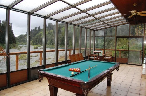 A sunroom with a pool table, overlooking a river and lush greenery through large glass windows.