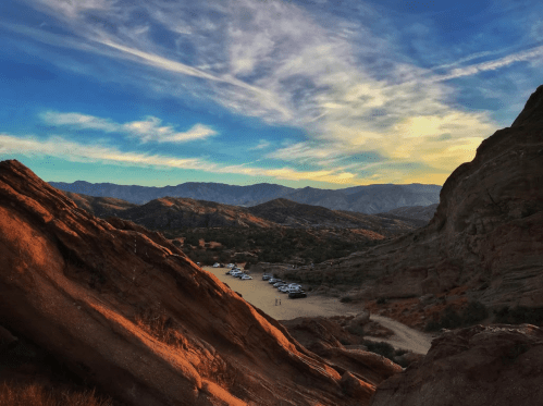 A scenic view of rocky terrain and mountains under a colorful sky, with a parking area visible in the distance.