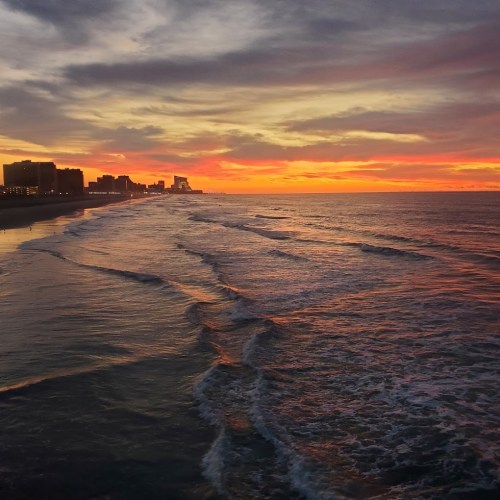 A serene beach at sunset, with gentle waves and a colorful sky reflecting on the water, and city silhouettes in the distance.