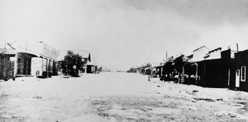 A deserted street in a historic town, lined with old buildings and a dusty road, captured in black and white.