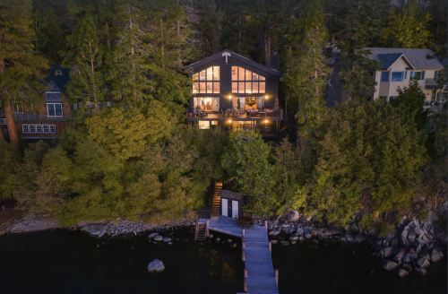 Aerial view of a modern cabin by a lake, surrounded by trees, with a wooden dock leading to the water.