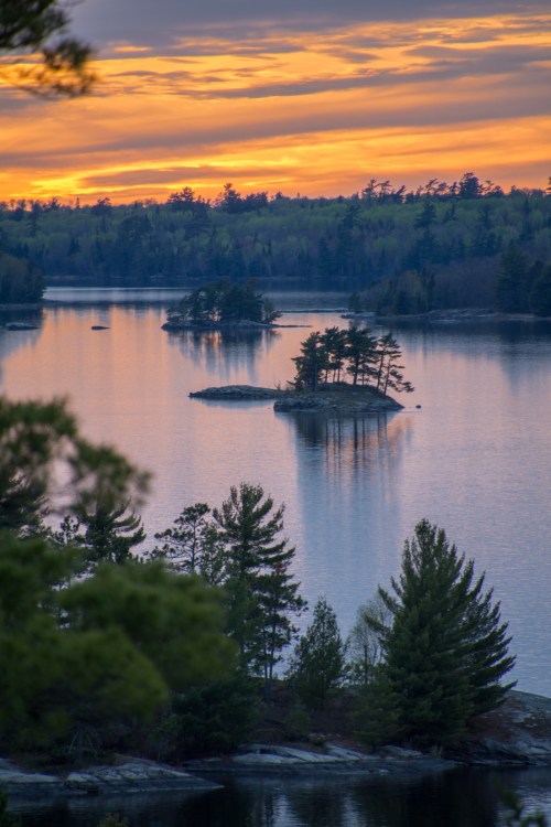 A serene lake at sunset, featuring small islands and trees reflecting on the water's surface.