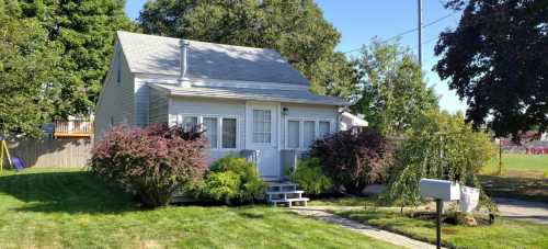 A small, charming house with a gray roof, surrounded by greenery and a well-maintained lawn on a sunny day.