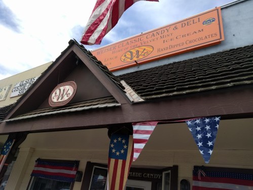 A storefront with an orange sign reading "We" and American flags hanging, featuring candy and deli items.