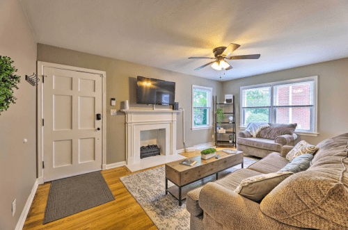 Cozy living room with a fireplace, TV, two sofas, and natural light from windows. Warm, inviting atmosphere.
