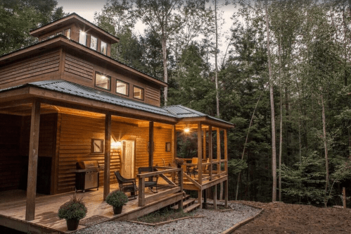 A modern wooden cabin with a porch, surrounded by trees in a serene forest setting.