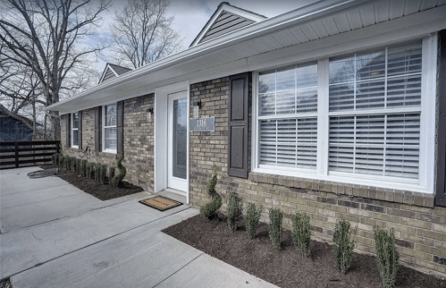A brick exterior of a house with a white door, windows with shutters, and neatly trimmed shrubs along the walkway.