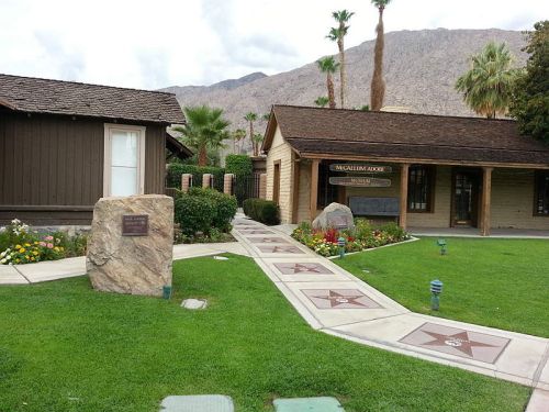 A pathway leads to a building with a sign, surrounded by greenery and mountains in the background.