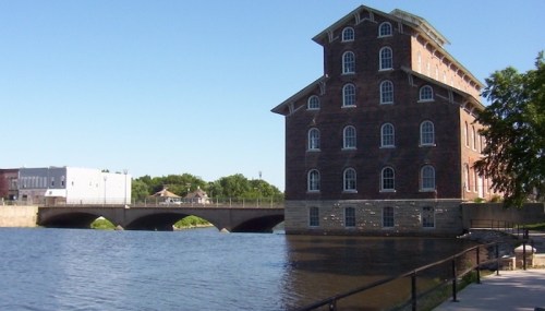 Historic brick building beside a river, with a bridge in the background and clear blue skies above.