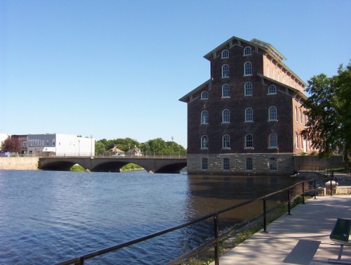 Historic brick building by a river, with a bridge in the background and a clear blue sky above.