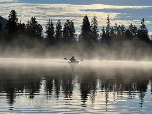 A person kayaking in a misty lake surrounded by trees, with soft clouds reflecting on the water's surface.