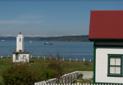 A scenic view of a calm bay with mountains in the background, featuring a lighthouse and a house with a red roof.