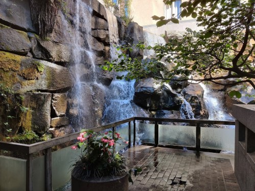 A serene waterfall cascades over rocks, surrounded by greenery and a small flower arrangement on a stone platform.