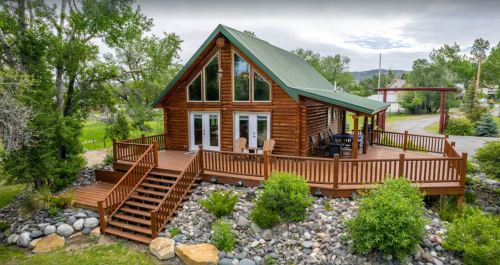 A charming log cabin with a green roof, large deck, and stone landscaping, surrounded by trees and greenery.