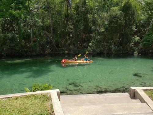 Two people kayak on a clear, green river surrounded by lush trees and vegetation. Steps lead down to the water.