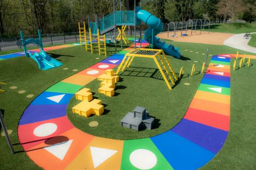 Colorful playground with slides, climbing structures, and a winding path featuring geometric shapes on the ground.