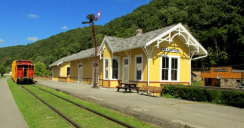 A yellow train station with a red train parked nearby, surrounded by green hills and trees.