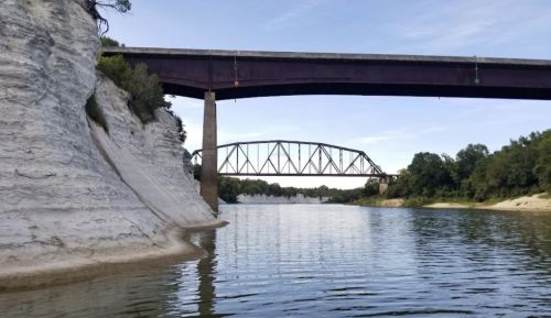 A river flows beneath two bridges, surrounded by white cliffs and green trees under a clear sky.