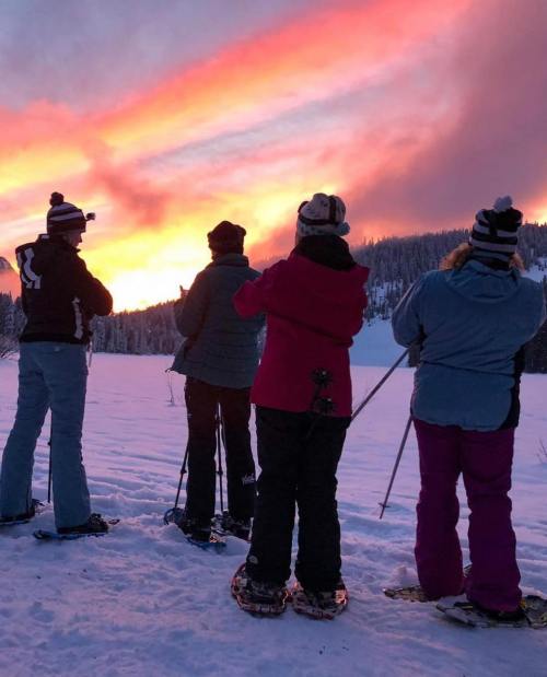 A group of four people in winter attire stands on snow, watching a vibrant sunset over a snowy landscape.