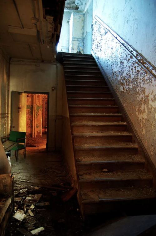 A dimly lit staircase leads upward, with peeling paint and debris scattered on the floor, hinting at abandonment.