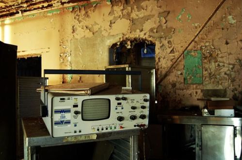 An old, dusty medical device sits on a metal cart in a dilapidated room with peeling walls.