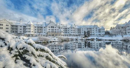 A snowy landscape featuring a large building reflected in a calm pond, surrounded by trees and a blue sky.