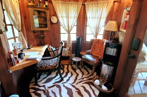 Cozy wooden room with a desk, chair, books, and a zebra-patterned rug, featuring large windows and warm lighting.