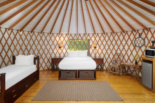 Cozy yurt interior featuring a double bed, single bed, wooden furniture, and a small kitchenette. Warm, inviting atmosphere.