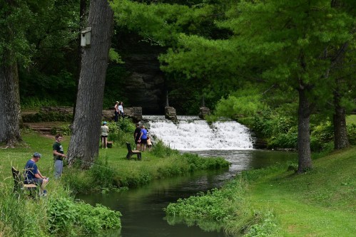 A serene park scene with a small waterfall, people fishing, and lush greenery along a winding stream.