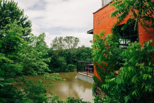 A vibrant red brick building surrounded by lush greenery, overlooking a calm, muddy river under a cloudy sky.