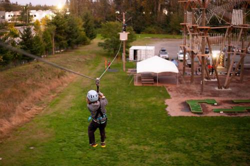 A child in a helmet zip lines through a park, with trees and an adventure course in the background.