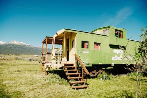 A green train caboose converted into a cozy cabin, set against a backdrop of mountains and clear blue skies.