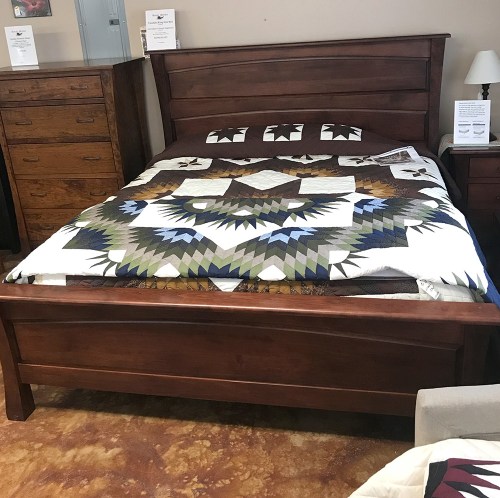 A wooden bed frame with a colorful quilt featuring geometric patterns, set in a furniture showroom.