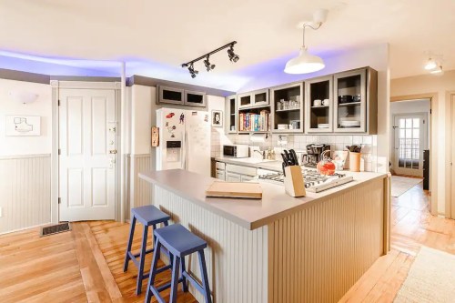 Modern kitchen with a central island, blue stools, and white cabinets, featuring warm wood floors and soft lighting.