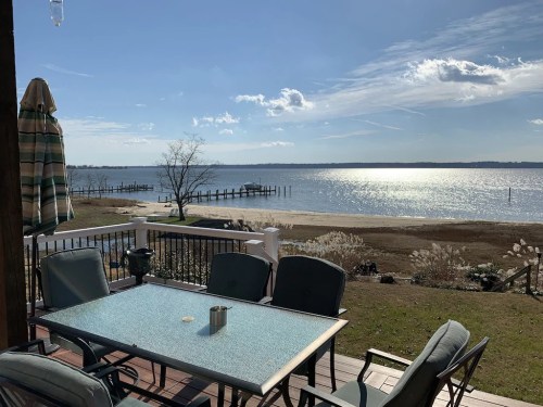 A serene view of a lake from a patio with a table and chairs, featuring a sandy shore and distant docks under a blue sky.