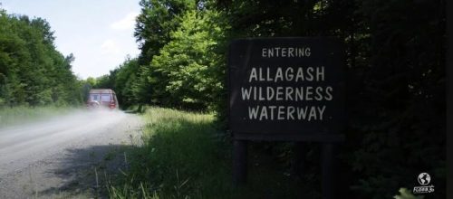 Sign marking the entrance to Allagash Wilderness Waterway, with a vehicle driving on a dirt road surrounded by greenery.