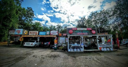 A vintage roadside shop with colorful signs, parked cars, and a cloudy sky in the background.