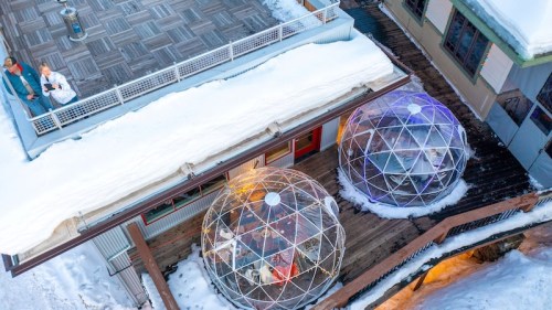 Two geodesic domes on a snowy deck, with people standing nearby, overlooking a winter landscape.
