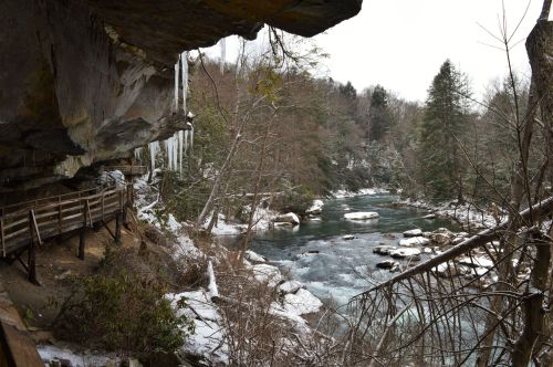 A snowy river scene with icicles hanging from a rock overhang and a wooden walkway along the water's edge.