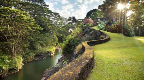 Lush green landscape with a winding stone path beside a tranquil river, framed by trees and sunlight.
